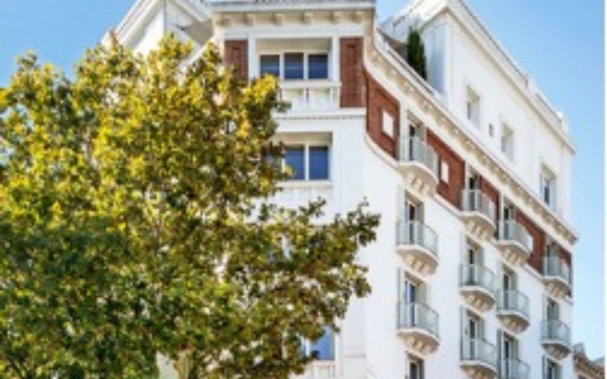 The white facade of a Madrid building with ornate balconies, partly hidden by a tree, under a blue sky.