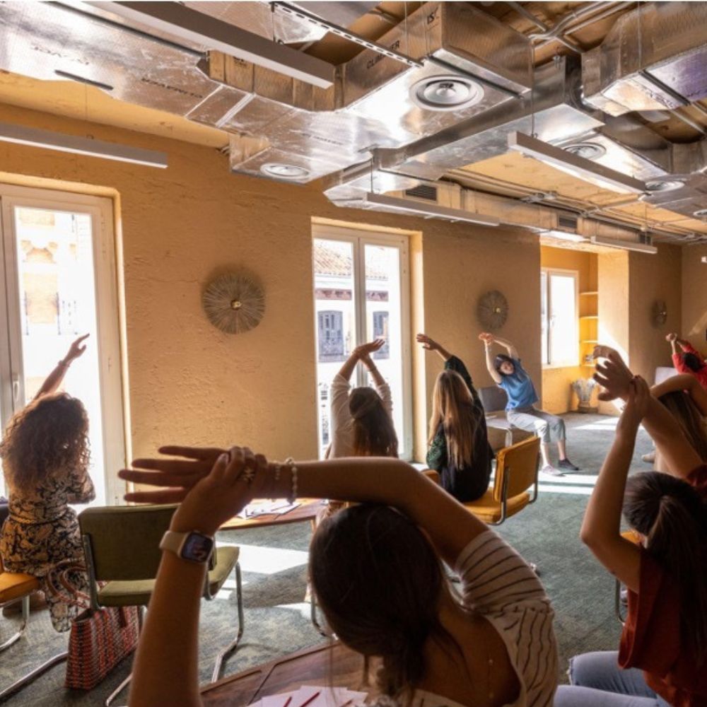 A dozen seated people stretch with their arms raised during a wellness session, in a room with ochre walls and exposed ventilation ducts.