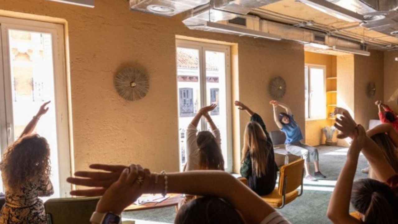 A dozen seated people stretch with their arms raised during a wellness session, in a room with ochre walls and exposed ventilation ducts.