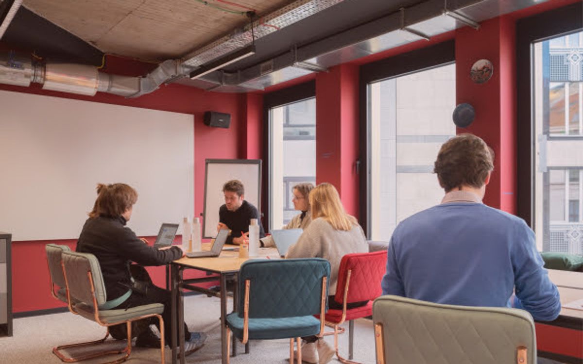 Five people gathered around a table with laptops in a red-walled room opening onto large windows.