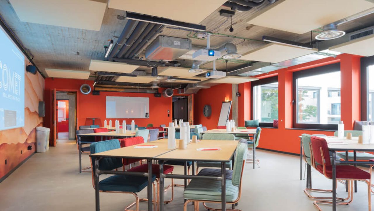 Vergaderzaal met feloranje muren, houten tafels en gekleurde stoelen, met het Comet-logo geprojecteerd op de achterwand.