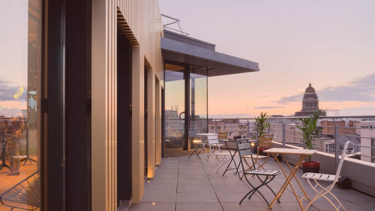 Terrasse en toiture aux tables de bistrot pliantes, au crépuscule, avec vue sur les toits de Bruxelles et le dôme du Palais de Justice.
