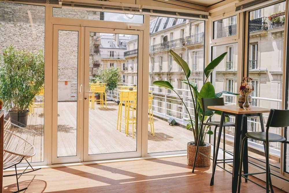 A bright conservatory opening onto a wooden terrace furnished with yellow tables and stools, facing Haussmann-style facades.