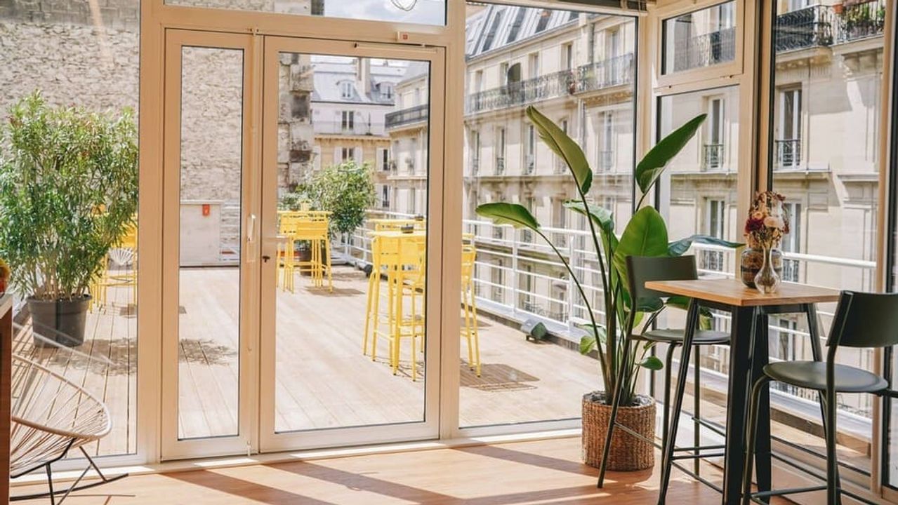 Une véranda lumineuse ouverte sur une terrasse en bois meublée de tables et de tabourets jaunes, face à des façades haussmanniennes.