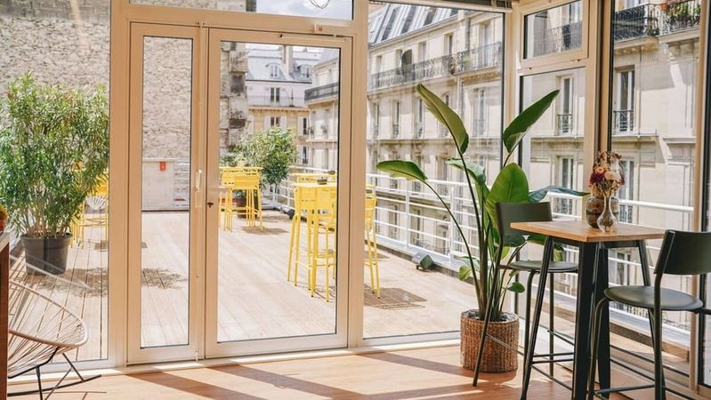A bright conservatory opening onto a wooden terrace furnished with yellow tables and stools, facing Haussmann-style facades.