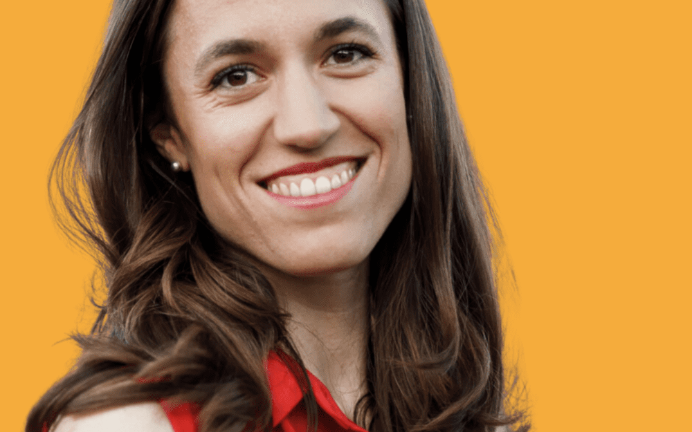 Romina Maillochaud, souriante, longs cheveux bruns et chemisier rouge sans manches, sur fond jaune.