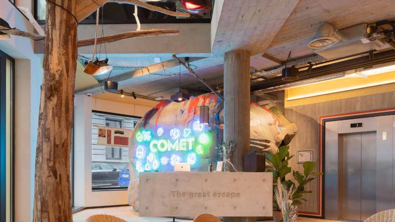 A reception hall crossed by a tree trunk, with a neon Comet sign on a rock-like backdrop and a concrete desk engraved "The great escape".