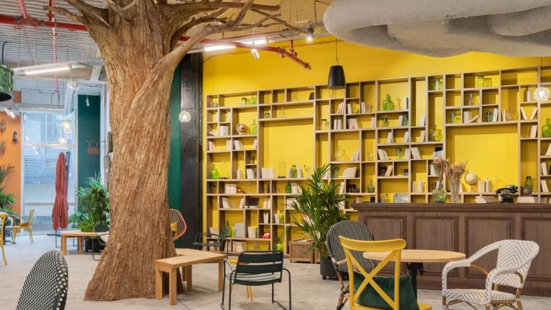 Een grote boom rijst op in een gemeenschappelijke ruimte met bistrostoelen, voor een felgele wand vol boekenkasten.