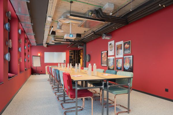A long meeting table lined with red and sea-green chairs in a room with red walls, with a whiteboard, a flipchart and a projector.