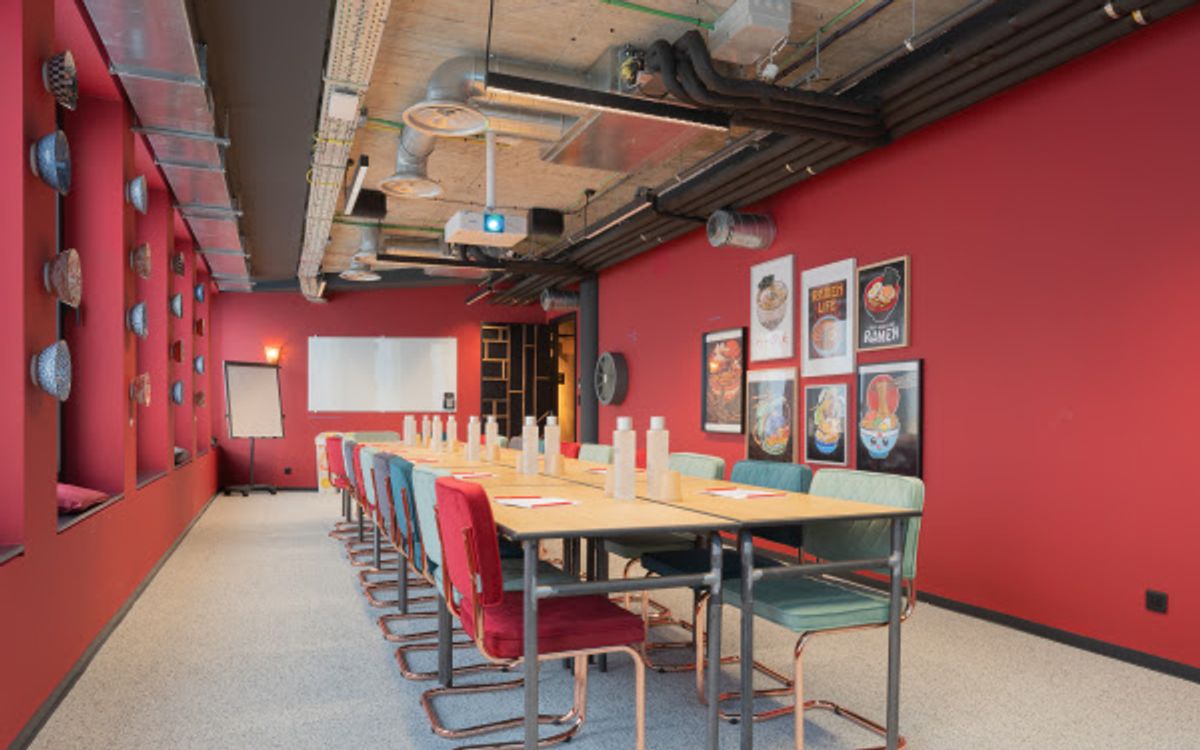 A long meeting table lined with red and sea-green chairs in a room with red walls, with a whiteboard, a flipchart and a projector.