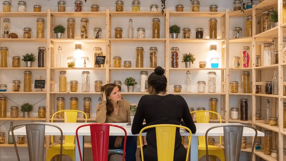 Twee collega's praten aan een lange witte tafel, in een ruimte met houten rekken vol glazen bokalen, onder industriële hanglampen.