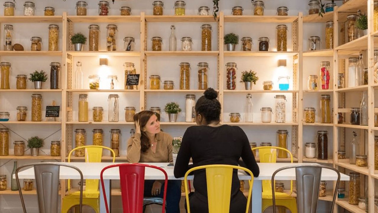 Two colleagues talk at a long white table in a room lined with wooden shelves full of glass jars, under industrial pendant lamps.