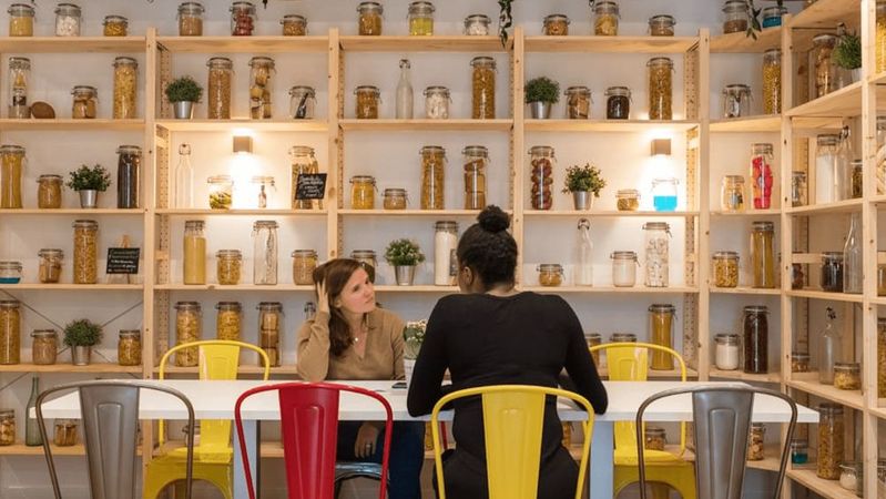 Two colleagues talk at a long white table in a room lined with wooden shelves full of glass jars, under industrial pendant lamps.
