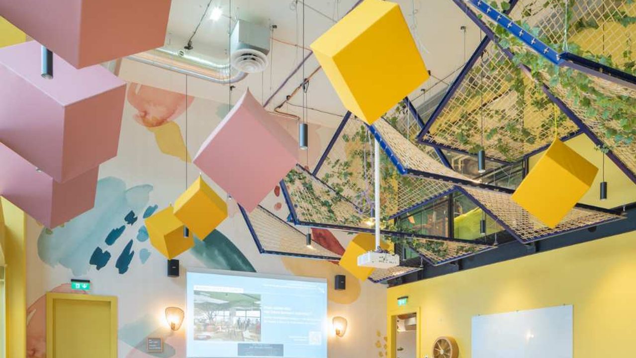 A bright, high-ceilinged meeting room with pink and yellow cubes hanging from the ceiling, a projection screen and long tables lined with colourful chairs.
