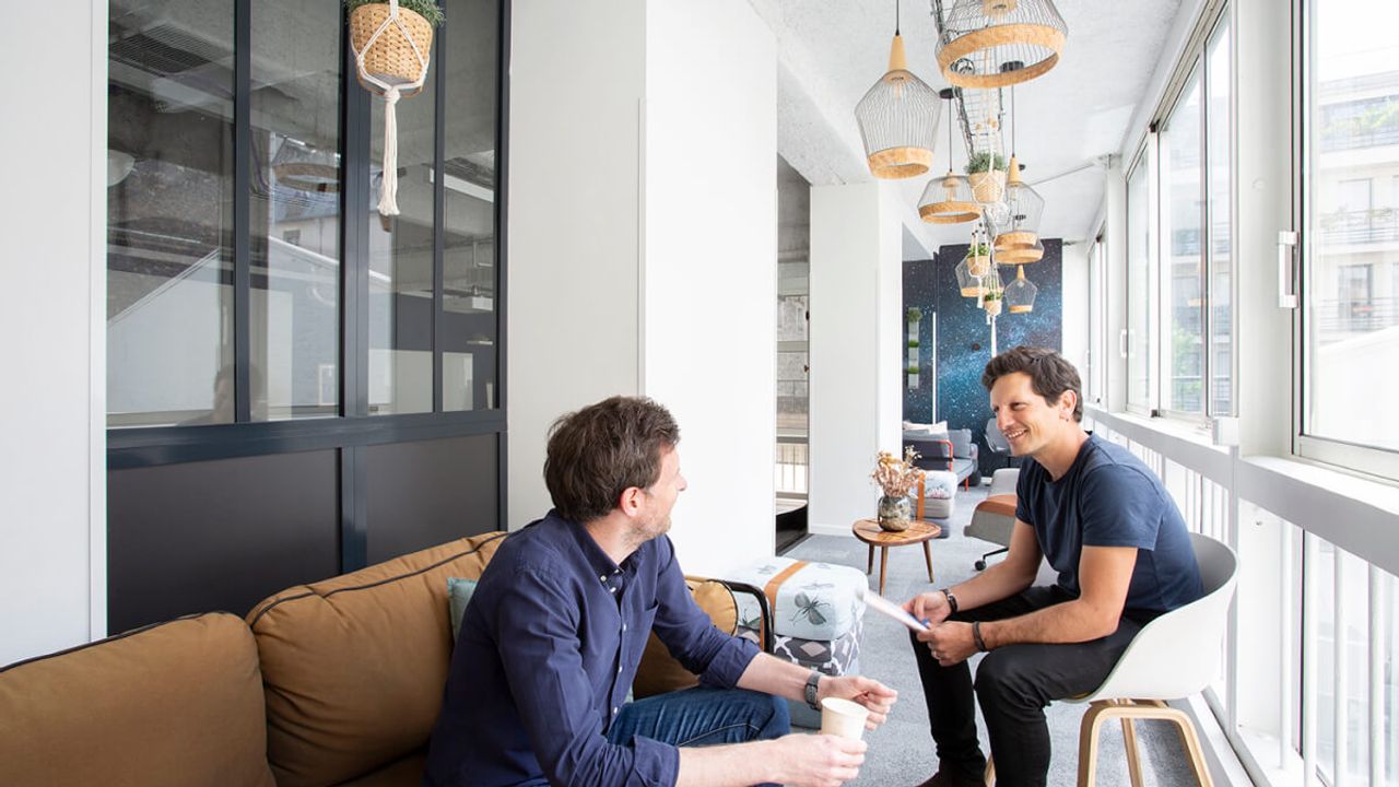 Two men sit facing each other in conversation in a bright lounge at a Comet venue, beneath rattan pendant lights and hanging plants.