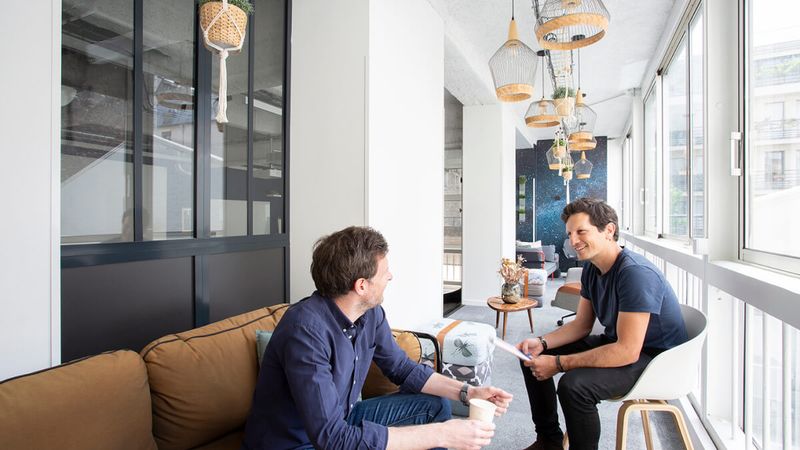 Deux hommes assis face à face échangent dans un salon lumineux d'un lieu Comet, sous des suspensions en rotin et des plantes suspendues.