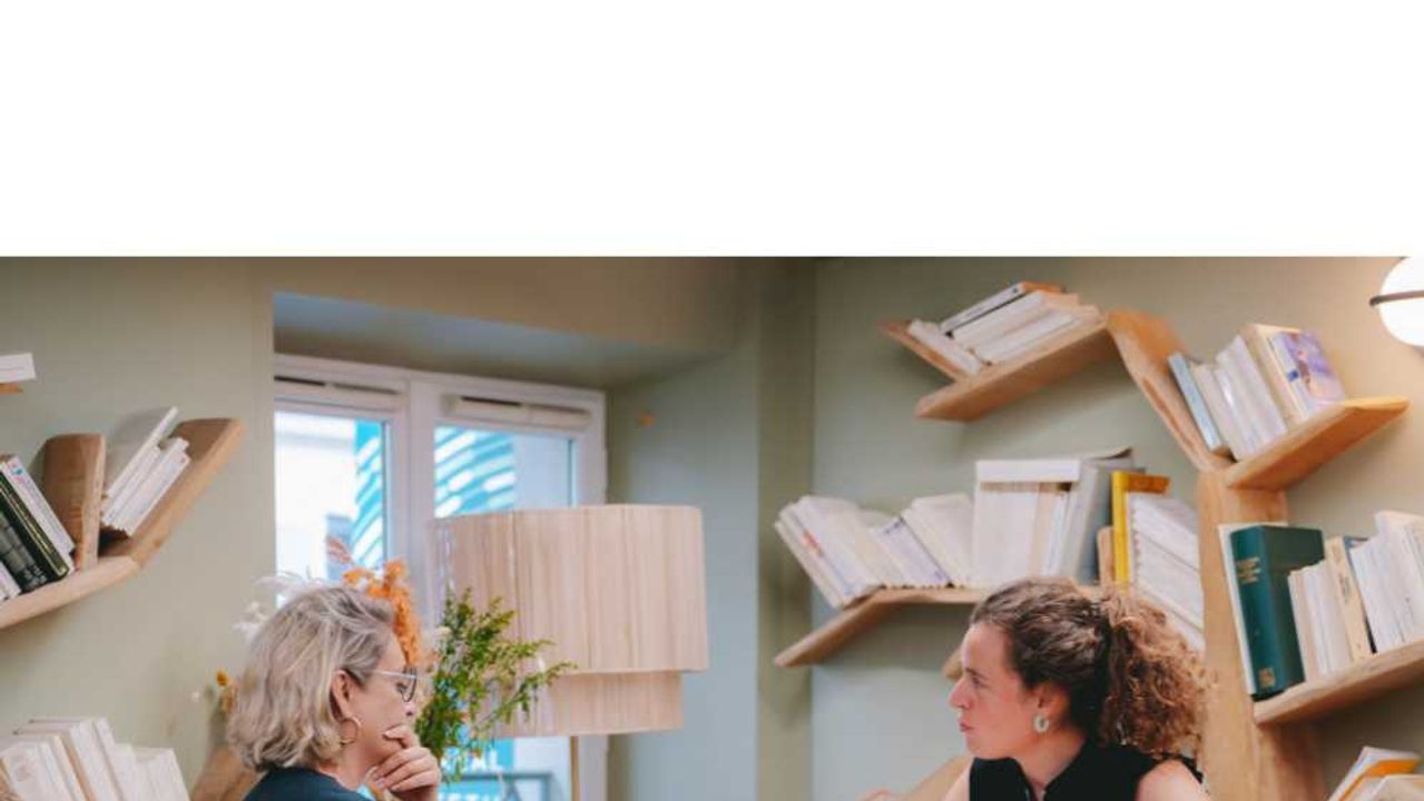 Two colleagues talk over their laptops in a Comet space lined with wooden shelves full of books.