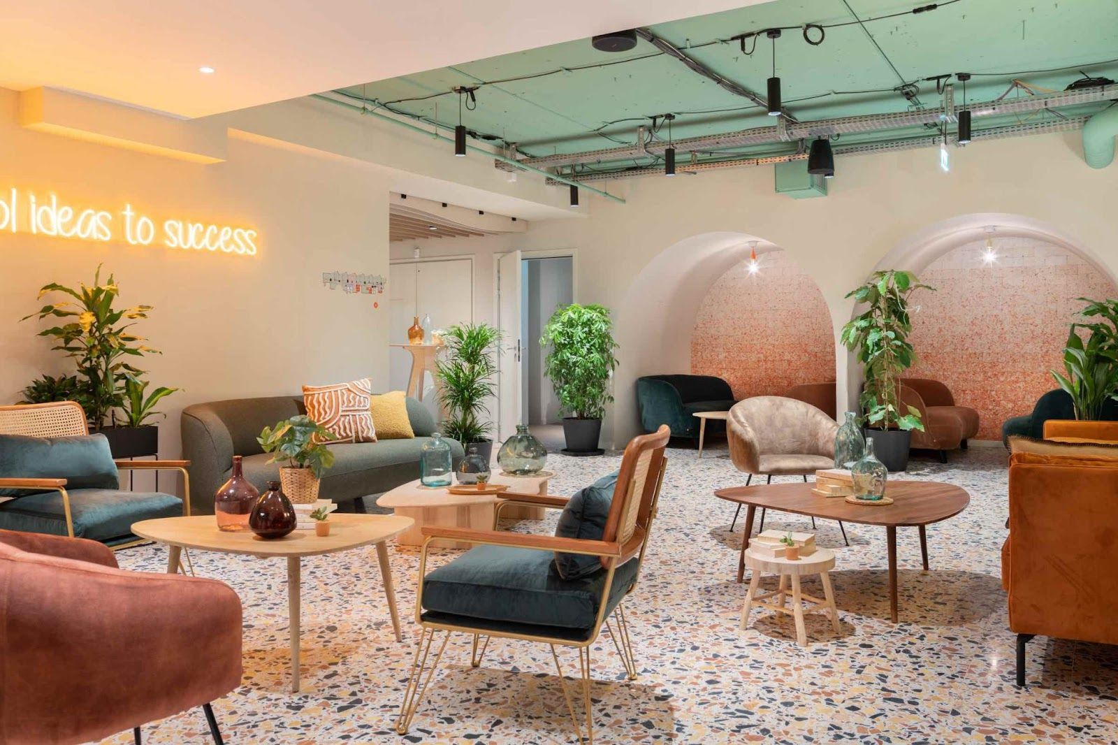 Un salón de Comet con butacas y sofás de colores, suelo de terrazo, plantas verdes y un rótulo de neón en la pared.