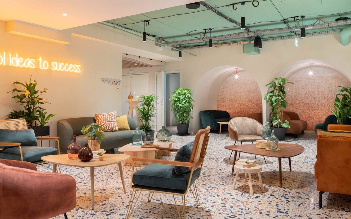 Un salon Comet aux fauteuils et canapés colorés, sol en terrazzo, plantes vertes et enseigne néon au mur.