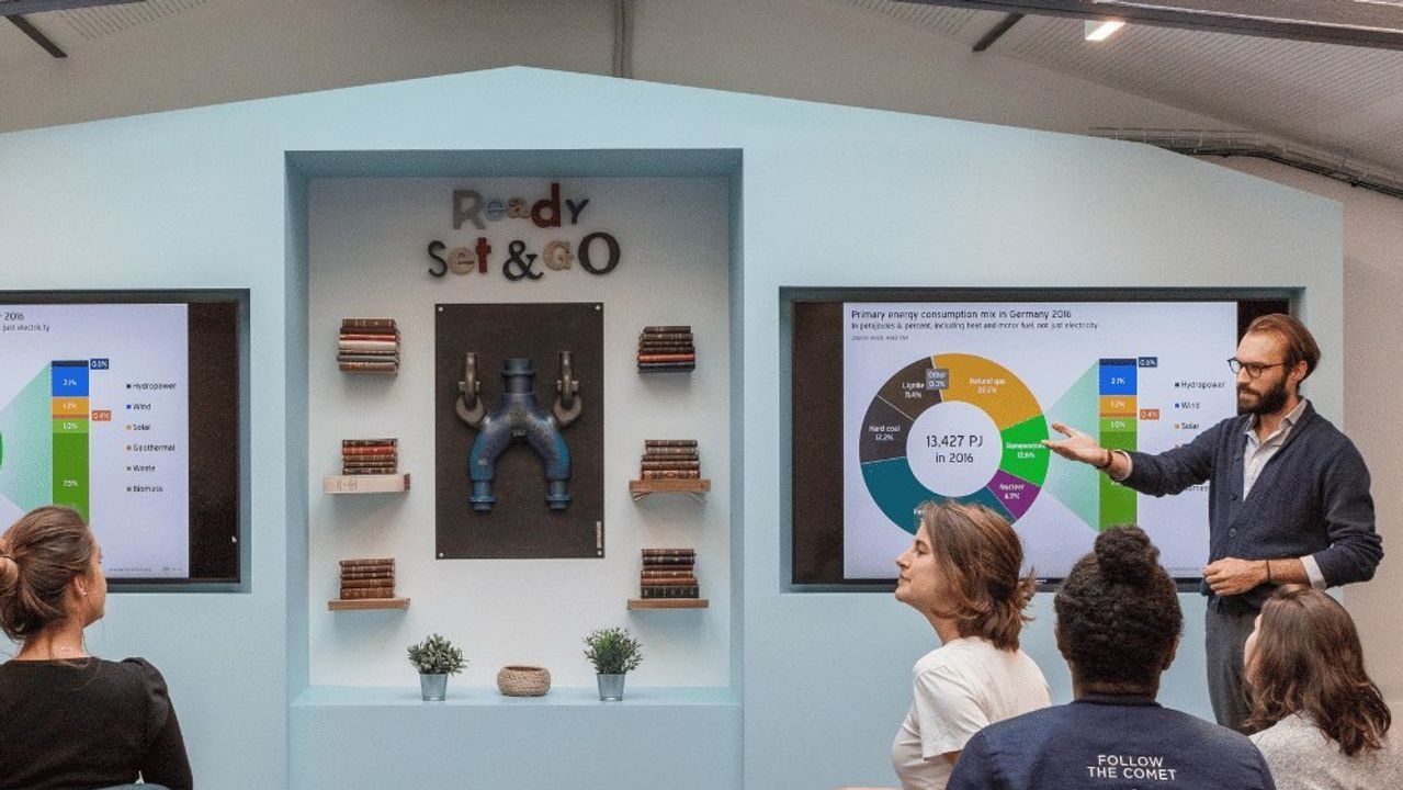 A speaker comments on a chart shown on screen to a small seated group, in a bright room topped by a glass roof and steel trusses.