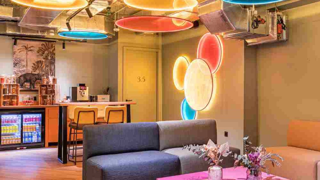 A lounge with large colourful illuminated discs hanging from the ceiling, a grey sofa, a bright pink coffee table and a drinks counter in the background.