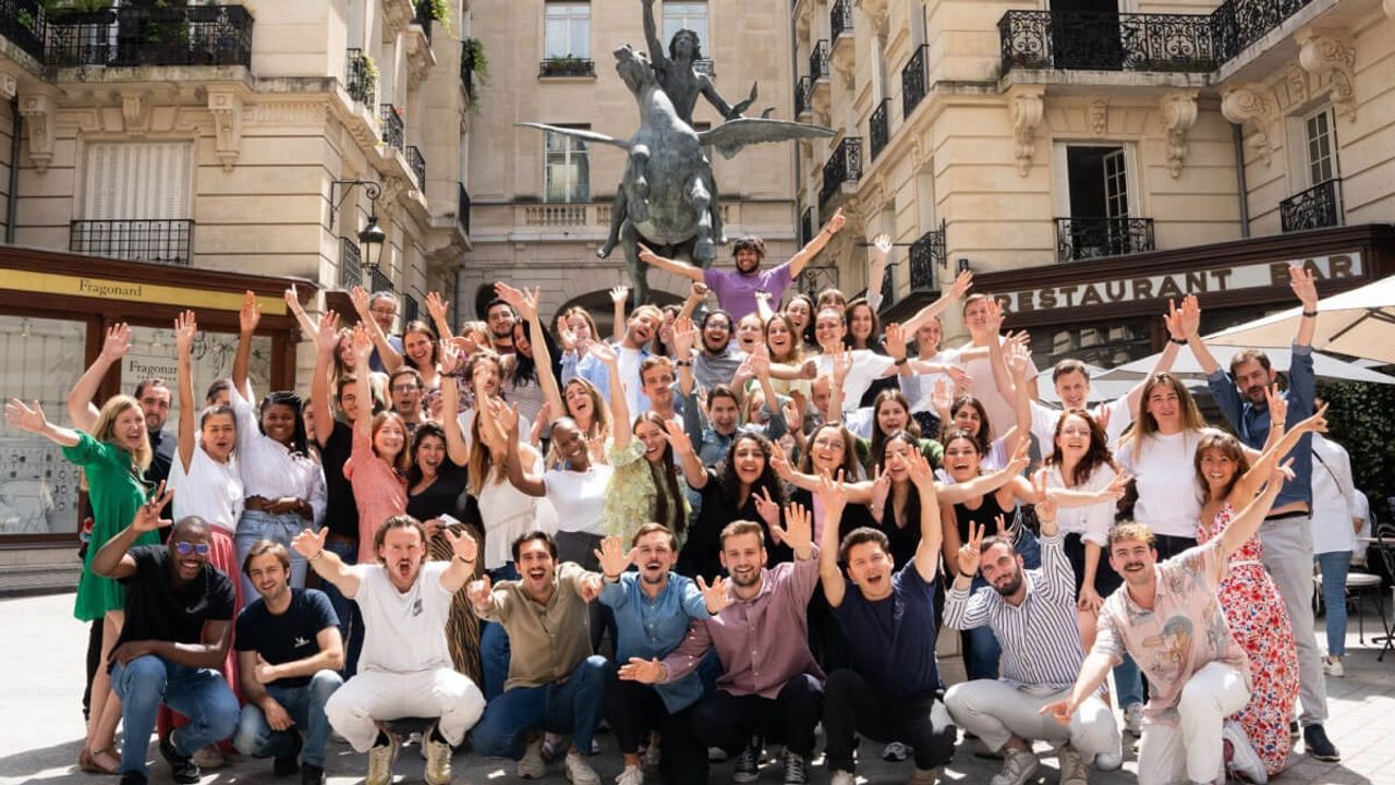 A large team posing with arms raised in a Paris square, in front of Haussmann buildings and an equestrian statue.