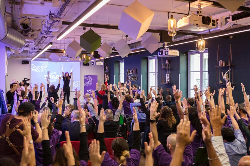 A packed room under purple lighting, where the audience raises their arms towards two speakers on stage.