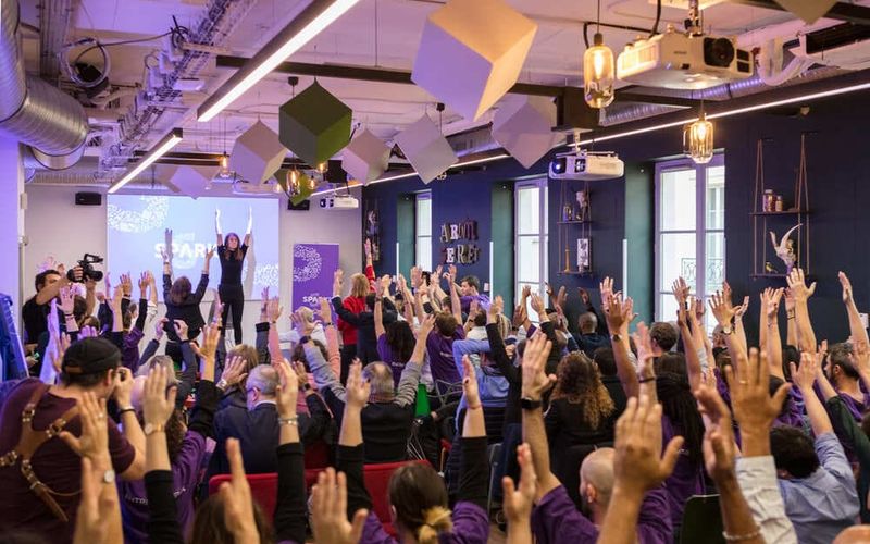 A packed room under purple lighting, where the audience raises their arms towards two speakers on stage.