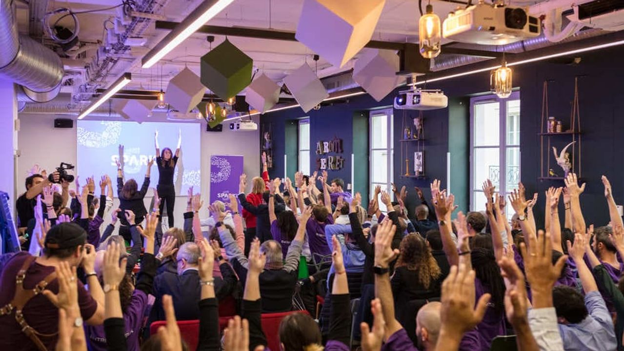 Een volle zaal in paars licht, waar het publiek de armen opsteekt naar twee sprekers op het podium.