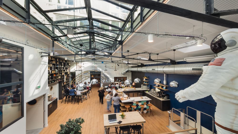 Une halle sous verrière métallique, avec bibliothèque murale, comptoir de restauration animé et une statue d'astronaute grandeur nature au premier plan.