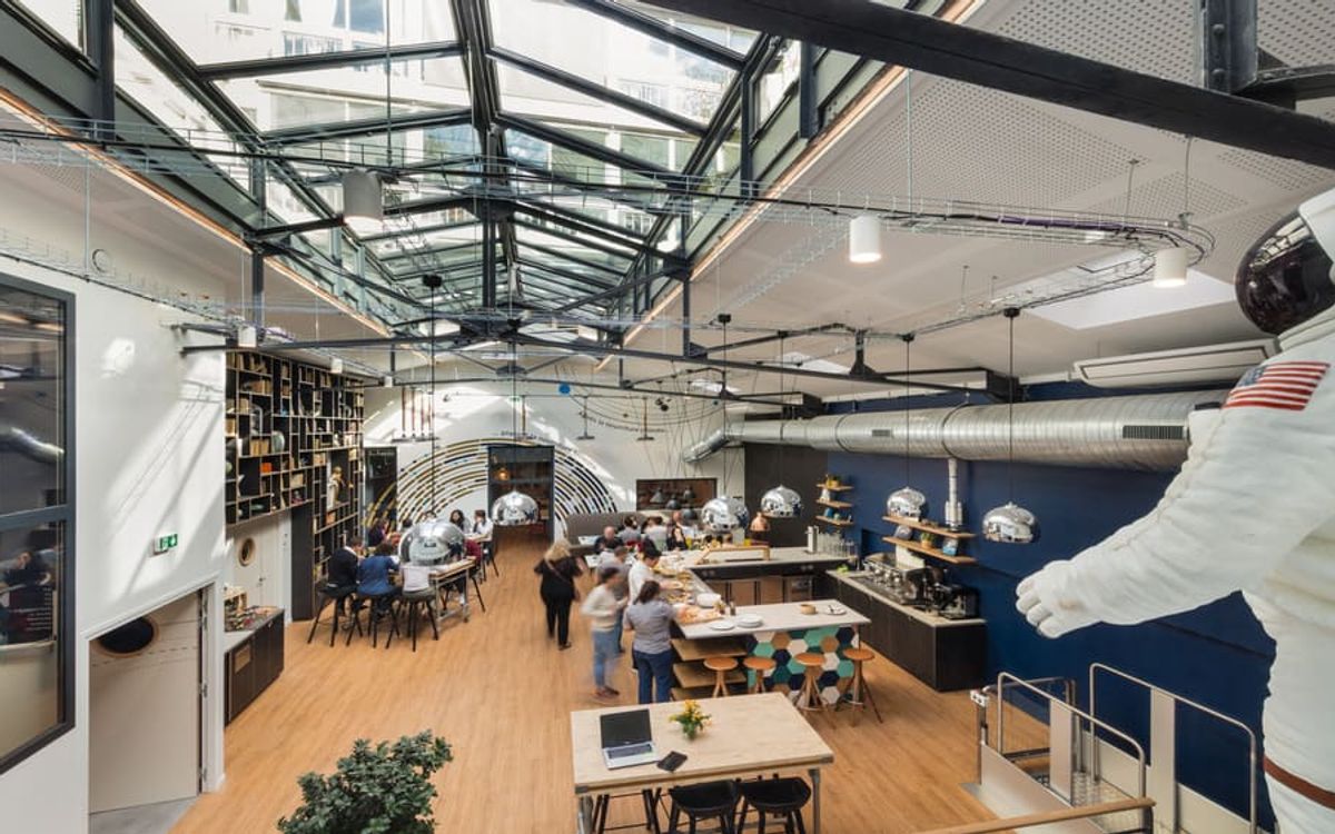 Une halle sous verrière métallique, avec bibliothèque murale, comptoir de restauration animé et une statue d'astronaute grandeur nature au premier plan.