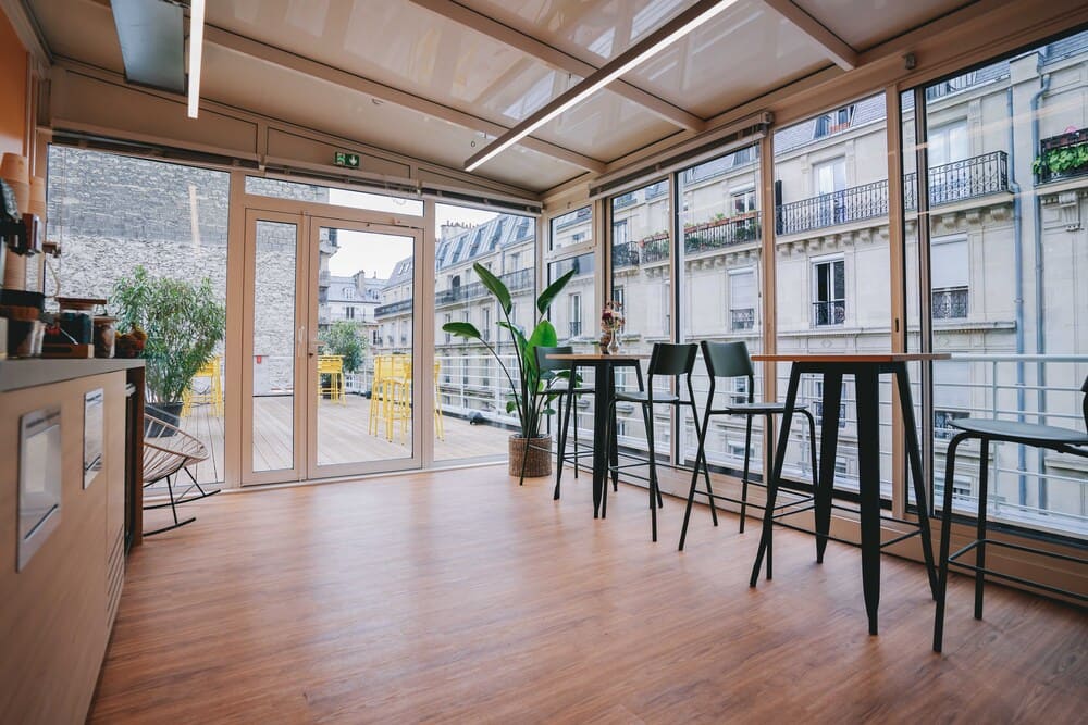 Un espace sous verrière meublé de mange-debout et de tabourets hauts, ouvert par de larges baies sur une terrasse en bois et les toits parisiens.