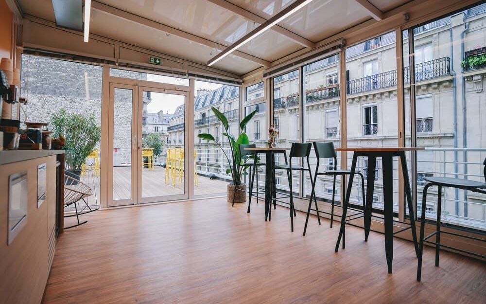A space under a glass roof furnished with high tables and bar stools, opening through wide bays onto a wooden terrace and the Paris rooftops.
