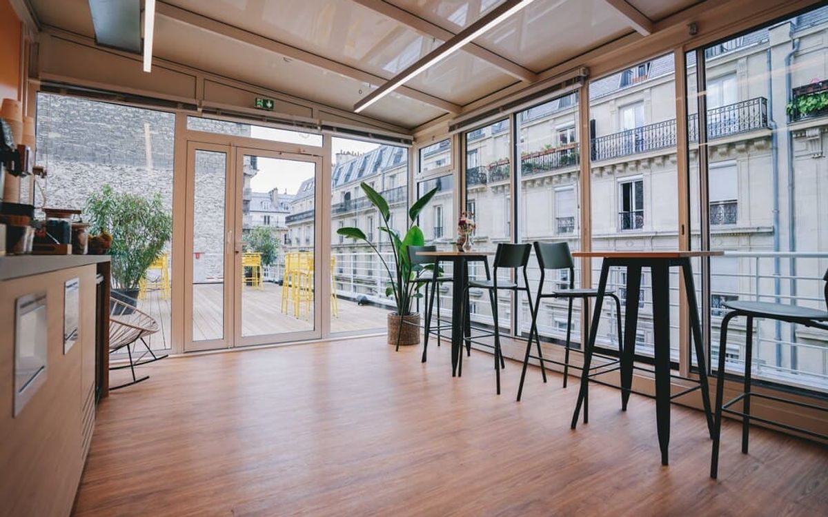Un espace sous verrière meublé de mange-debout et de tabourets hauts, ouvert par de larges baies sur une terrasse en bois et les toits parisiens.