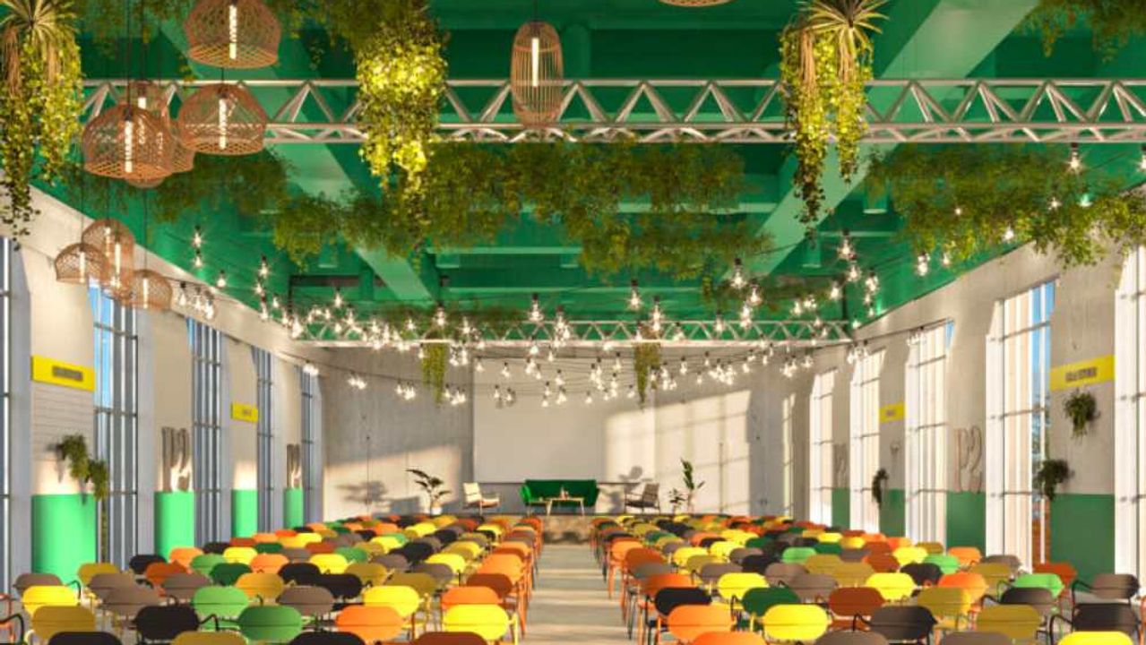A large event hall with green-painted beams, hanging plants and rattan pendant lights, where rows of colourful chairs face a small stage.