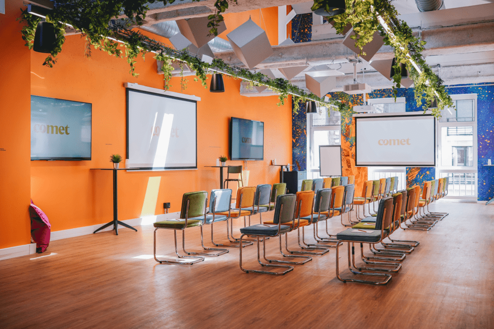 Une salle aux murs orange disposée en format conférence, avec des rangées de chaises colorées face à trois écrans, sous des guirlandes de feuillage.