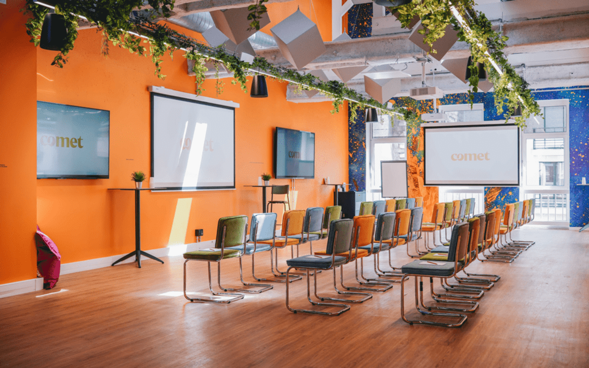 Une salle aux murs orange disposée en format conférence, avec des rangées de chaises colorées face à trois écrans, sous des guirlandes de feuillage.