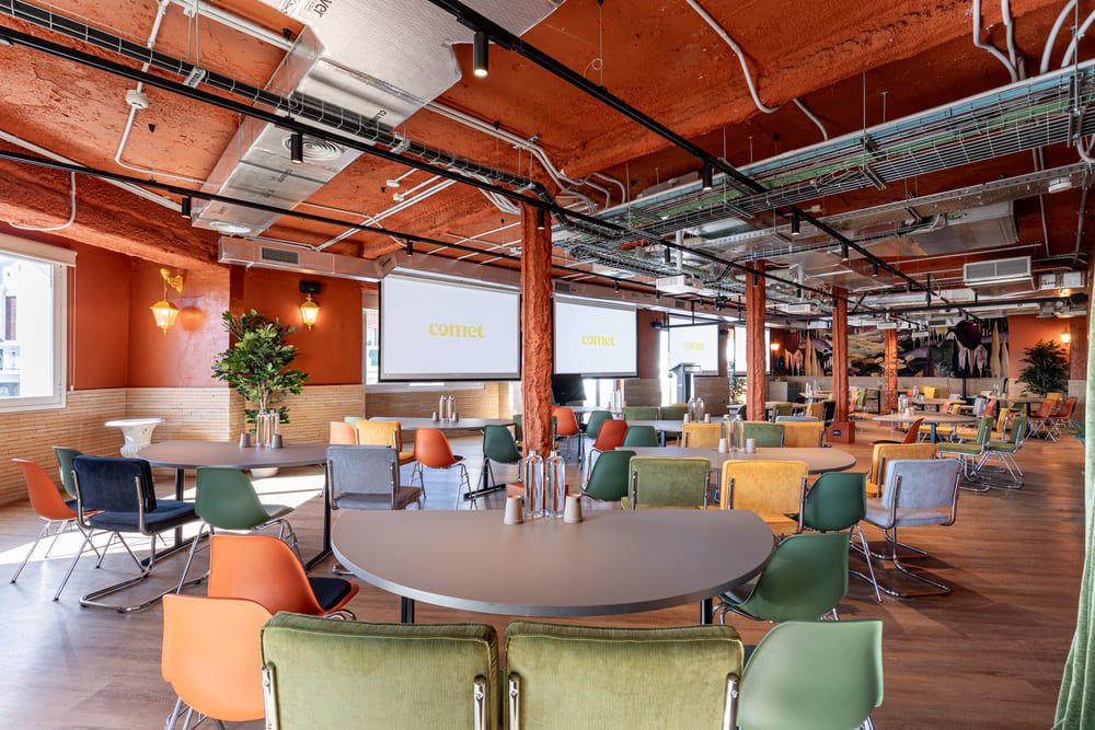 A vast open floor with orange beams and columns, laid out with round tables and colourful chairs facing two projection screens.