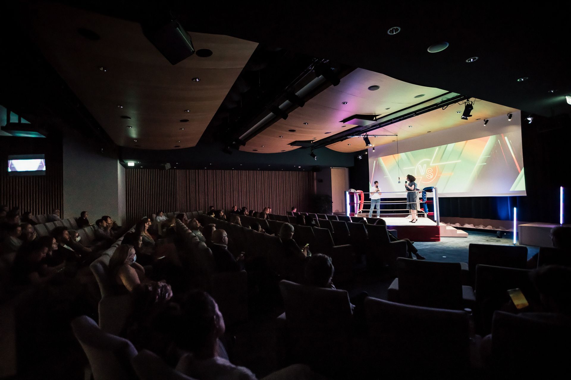 Wide view of the dimly lit auditorium: the audience, seated in armchairs, faces two speakers in the lit boxing ring.