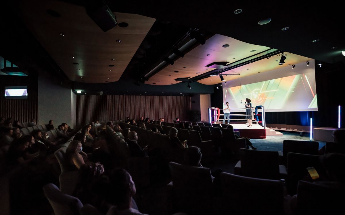 Wide view of the dimly lit auditorium: the audience, seated in armchairs, faces two speakers in the lit boxing ring.