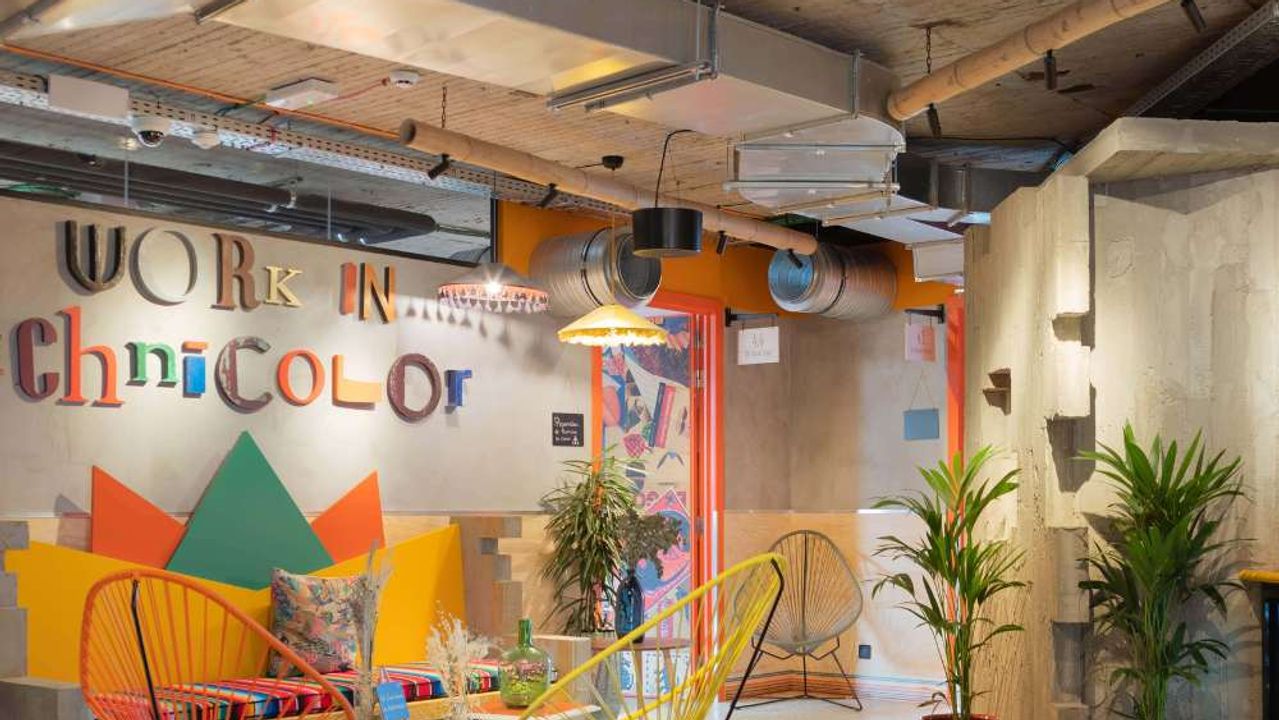 Un salón colorido con sillas Acapulco naranja y amarilla bajo un techo de bambú y hormigón visto, frente a una pared con el rótulo «Work in Technicolor».