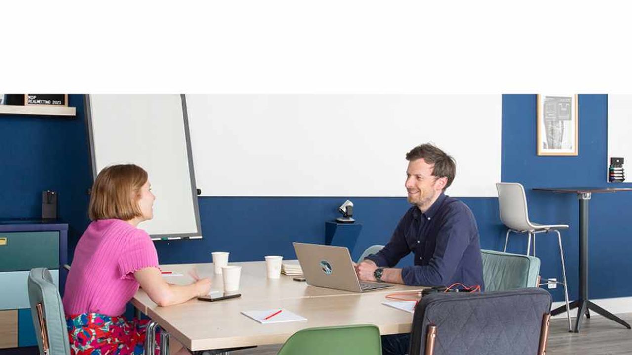 Two colleagues talk across a meeting table, in front of a blue wall and a flip chart, with an open laptop between them.