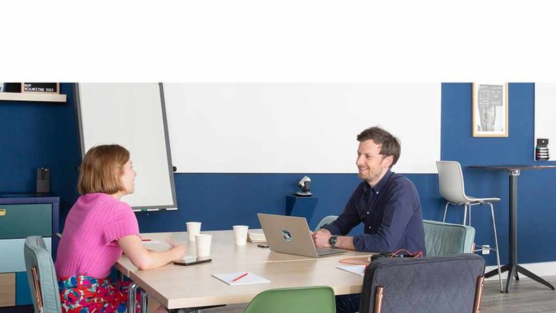 Two colleagues talk across a meeting table, in front of a blue wall and a flip chart, with an open laptop between them.