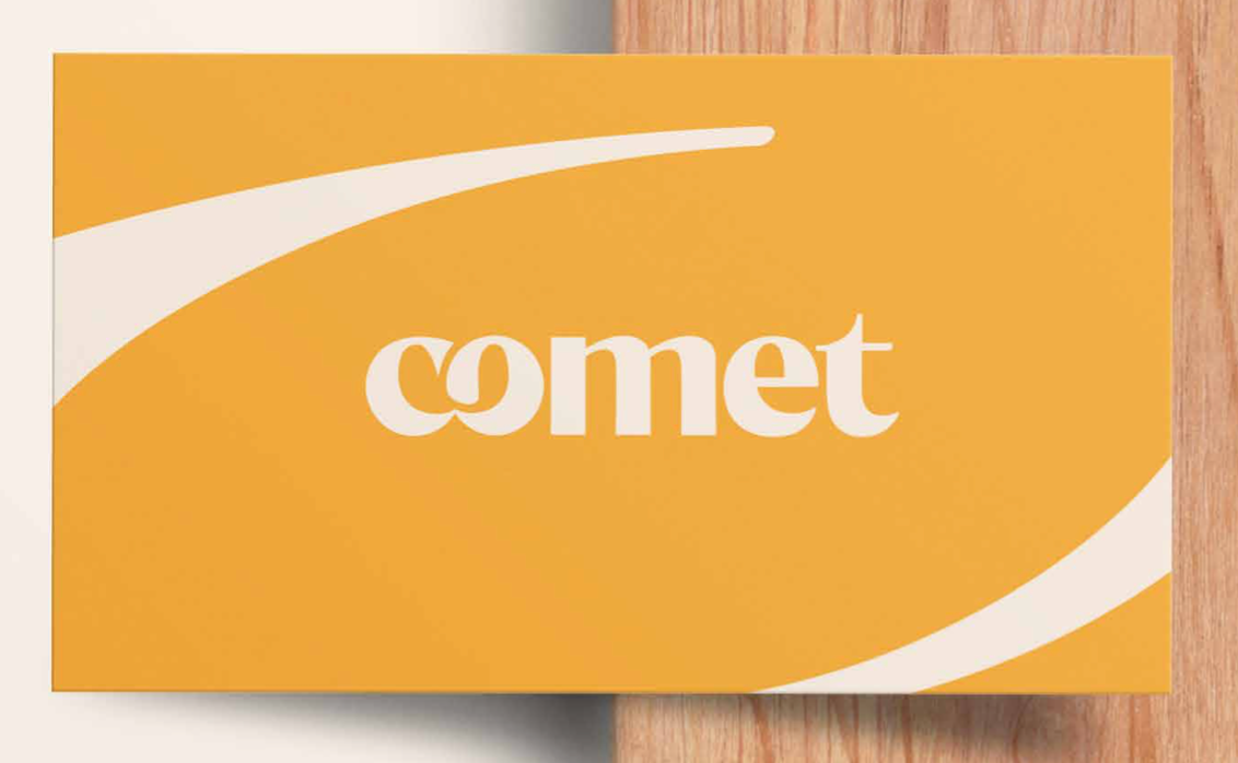 Una tarjeta de visita amarilla con el nuevo logotipo de Comet de letras redondeadas, sobre una superficie de madera clara.