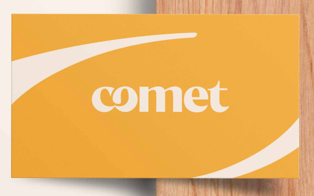 Une carte de visite jaune portant le nouveau logotype Comet aux lettres arrondies, posée sur un plan de travail en bois clair.