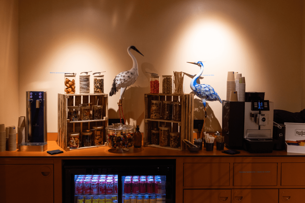 Un comptoir en libre-service avec machine à café, fontaine à eau, bocaux de snacks et deux hérons décoratifs en papier.