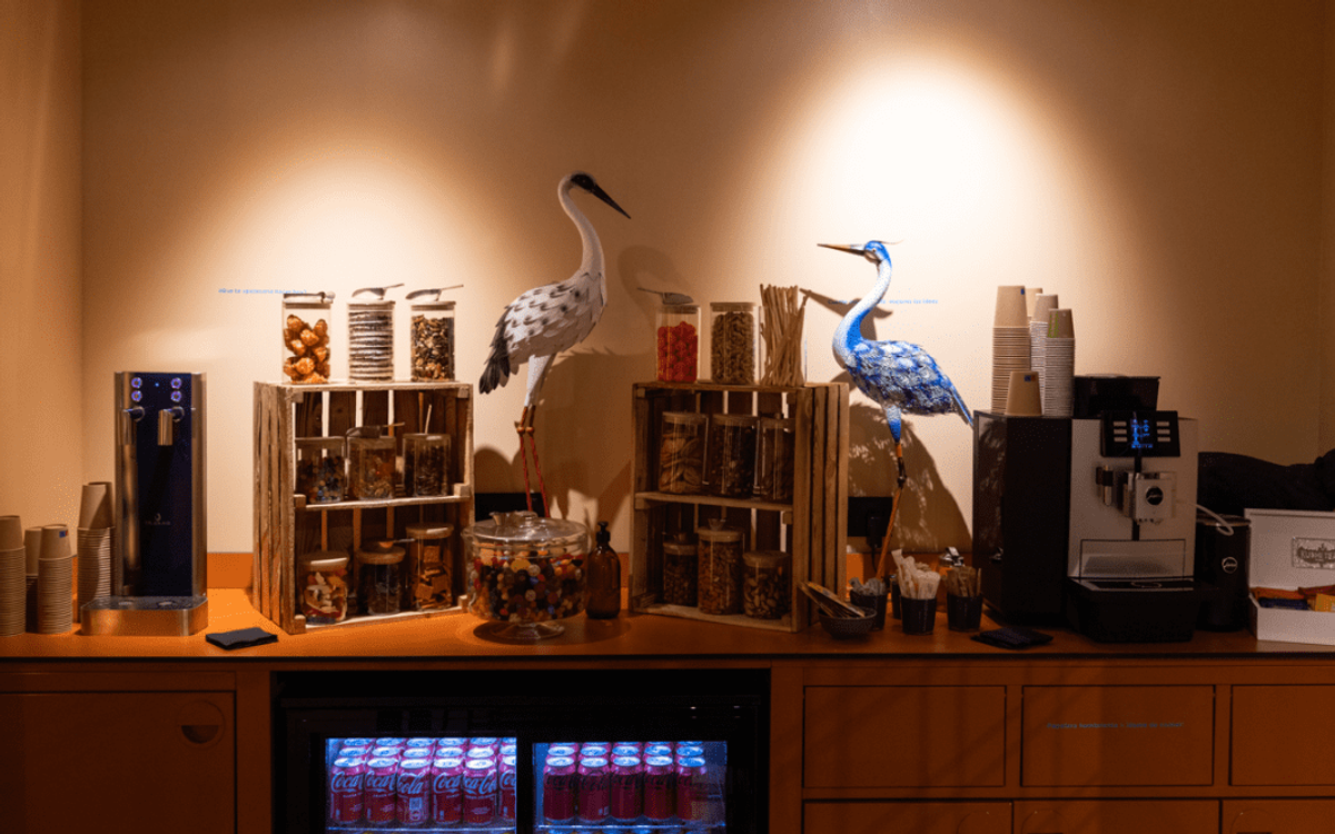 Un comptoir en libre-service avec machine à café, fontaine à eau, bocaux de snacks et deux hérons décoratifs en papier.