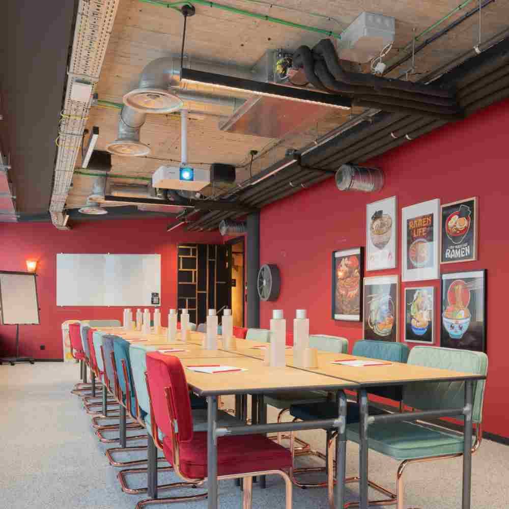 A meeting room with red walls, a long pale table lined with red and sea-green vintage chairs, under a wooden ceiling with exposed ducting.