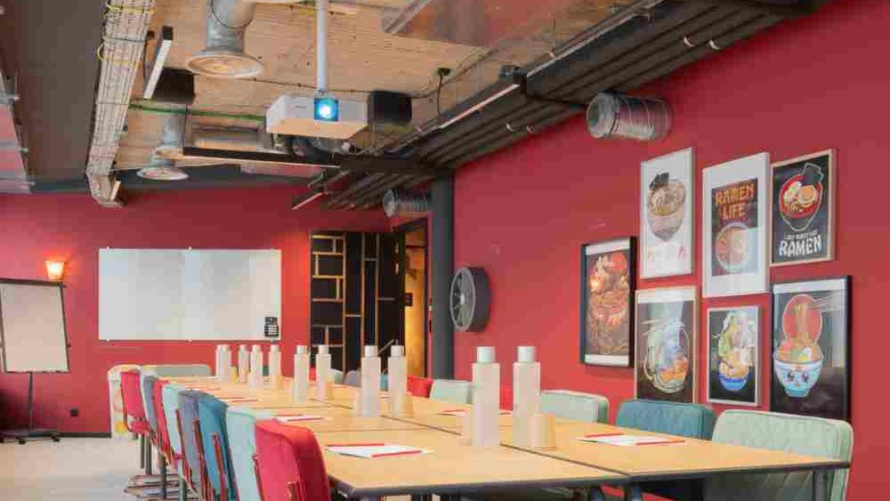 A meeting room with red walls, a long pale table lined with red and sea-green vintage chairs, under a wooden ceiling with exposed ducting.