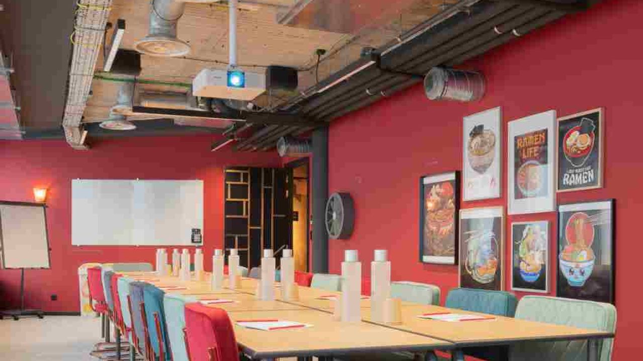 A meeting room with red walls, a long pale table lined with red and sea-green vintage chairs, under a wooden ceiling with exposed ducting.